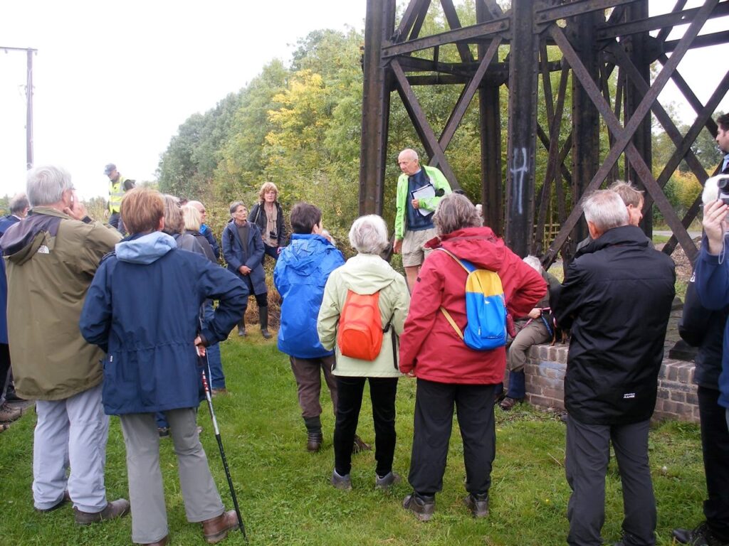 The image shows a group of people on a guided tour under the viaduct. They are being informed of about the design of the viaduct by a guide from the Friends of Bennerley Viaduct.